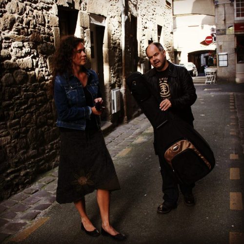 Andy and Viil with a guitar bag in an old cobbled street partly in sunlight.